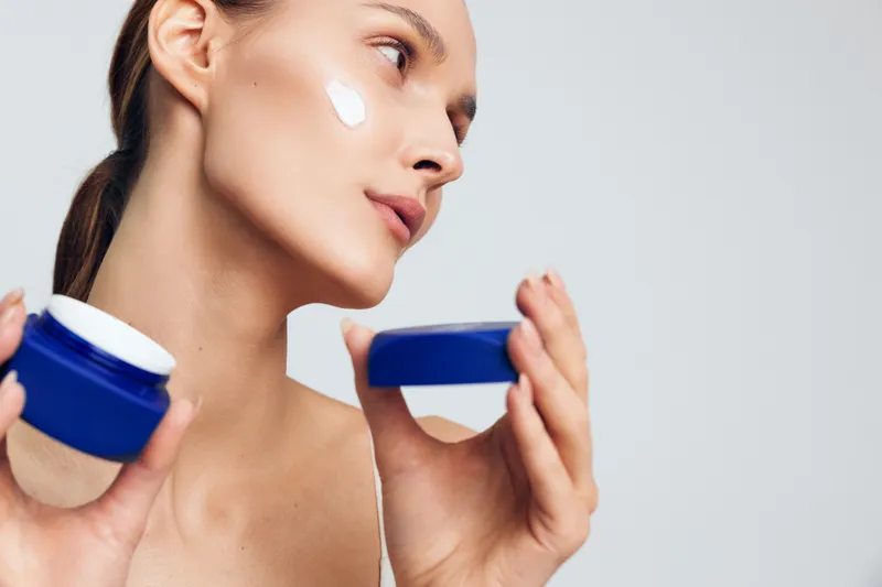 A young woman applies white face cream on her cheek holding a blue jar and lid. Skincare concept with natural beauty and healthy skin on a light background.