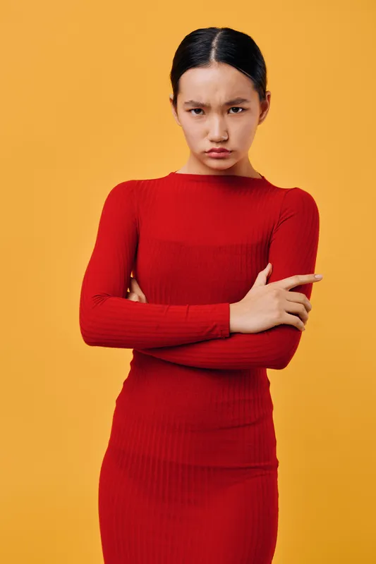Woman in red dress with crossed arms and serious expression standing against yellow background. Young adult female posing with intense look and sleek hairstyle in studio lighting.