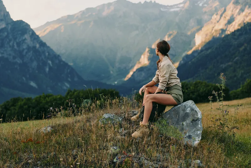 Bright outdoor lifestyle scene with a woman sitting on a rock in a meadow, mountains in the background, relaxed pose, natural light, gentle breeze, warm tones