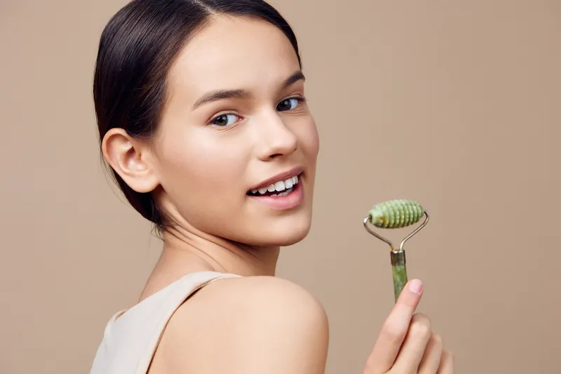 Young woman of Asian descent smiling while using a jade facial roller, emphasizing beauty and skincare routines with a soft beige background