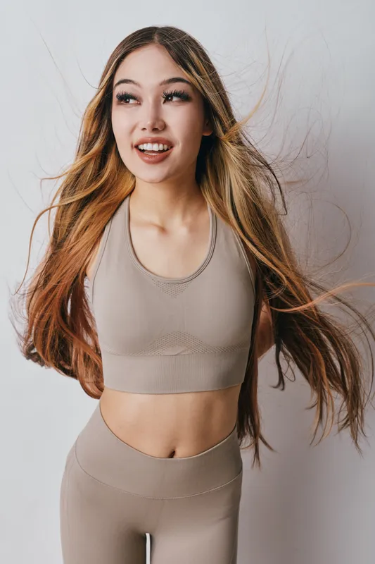 Young woman with long hair in sportswear smiling and looking away on white background. Fitness model in neutral outfit posing with flowing hair for healthy lifestyle.
