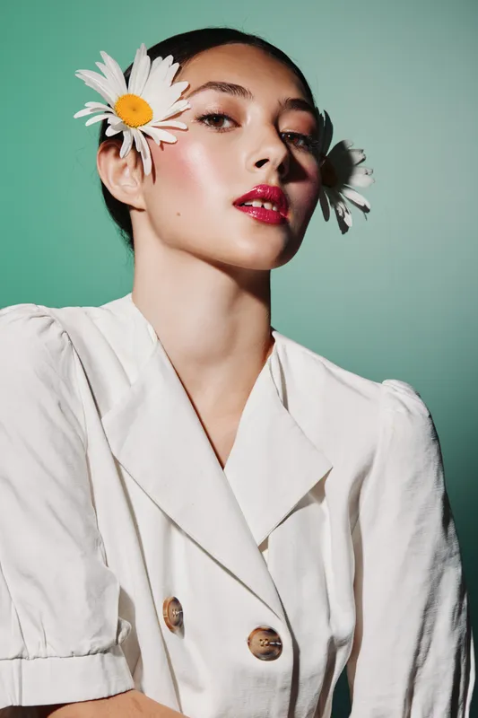 Young woman with bright red lipstick and highlighter makeup wears white jacket with daisy flowers in her hair. Clean modern beauty and fashion creative on green background.