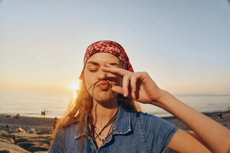 lifestyle woman in modern boho-western denim street style with a red bandana posing playfully at sunset by the beach, film color tones capturing warm, relaxed vibes