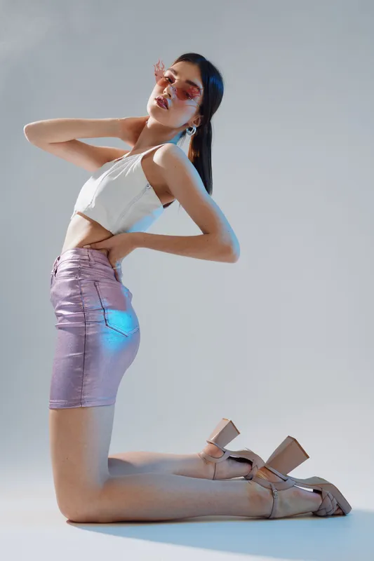 Young woman in a white top and shiny pink skirt poses kneeling with eyes closed and hand on neck in studio. Concept of beauty, fashion, elegance, and modern style.