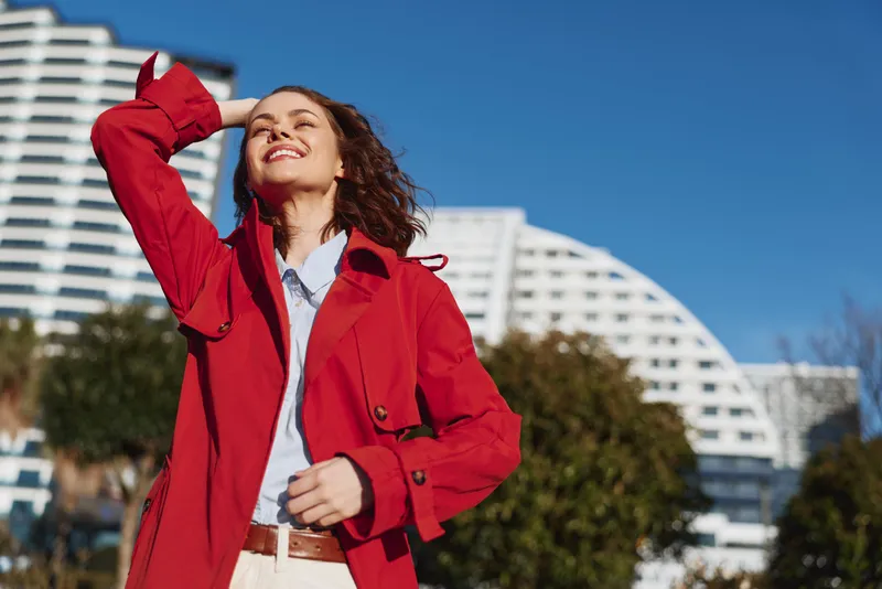 Young woman with curly hair smiles joyfully while enjoying a sunny day outdoors wearing a red coat and light blue shirt against a modern urban background.