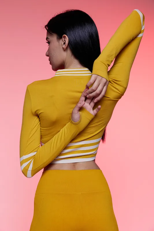 Woman sport fitness stretching her arm behind back wearing mustard yellow activewear against pink background, demonstrating flexibility and workout preparation.