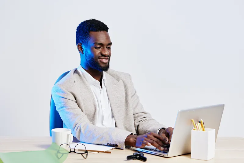 Smiling young man working on laptop in modern office setting, casual office attire, using computer, sitting at desk with coffee mug, glasses, and stationery, bright and professional environment, conce