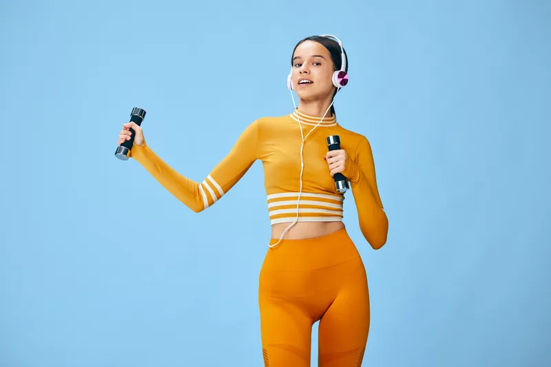 Young woman fitness portrait in orange activewear holding dumbbells and wearing headphones, upbeat sport concept in a clean blue studio background with full body motion.