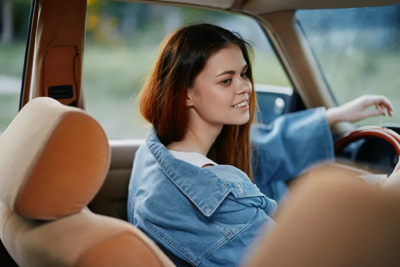 Young woman smiling in a car with a denim jacket, capturing a carefree and joyful emotion, with soft natural light filtering through the window.
