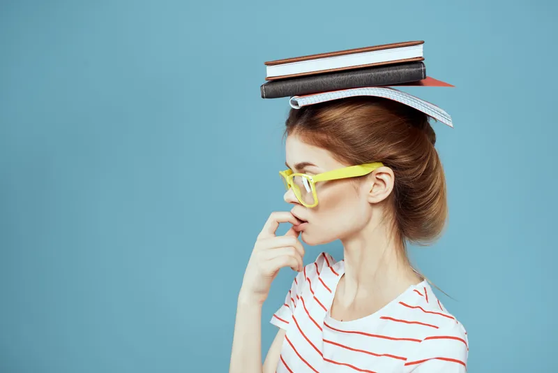Студентка держит книги на голове и в жёлтых очках на синем фоне, обрезанный вид