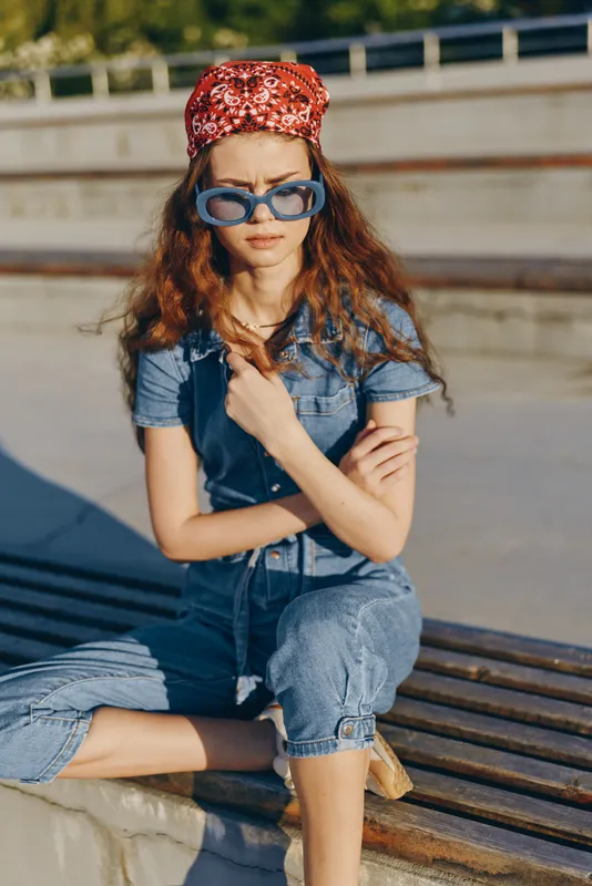 young woman wearing sunglasses and a red bandana sits outdoors on a sunny day in casual denim clothing and looks thoughtful in summer light