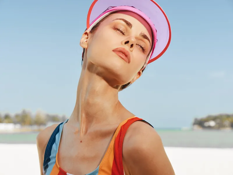 Closeup of a sunlit woman on a bright beach, wearing a colorful swimsuit and pink visor, eyes closed, savoring warm breeze and radiant summer glow.
