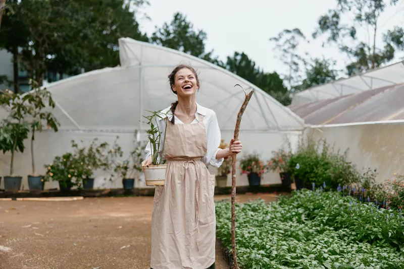 Happy woman with a plant in a greenhouse, wearing a light colored apron and holding a stick, surrounded by greenery, portraying a joyful gardening concept