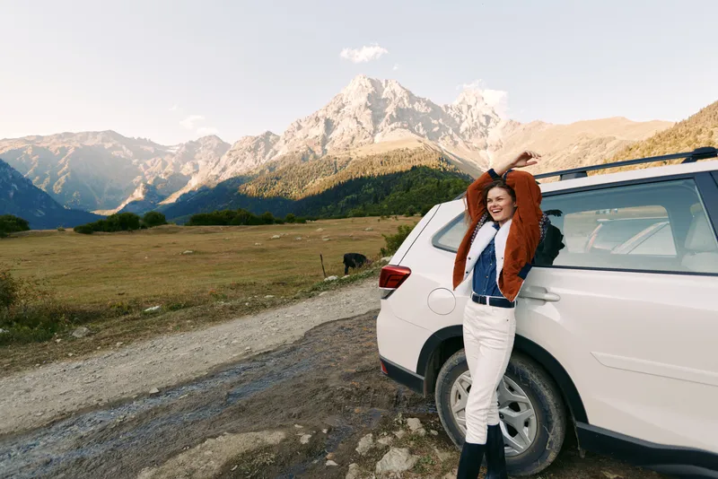 Woman by car in mountain landscape, smiling with arms raised beside white SUV on a gravel road, travel adventure in an open meadow with scenic peaks, clear sky and relaxed vibe.