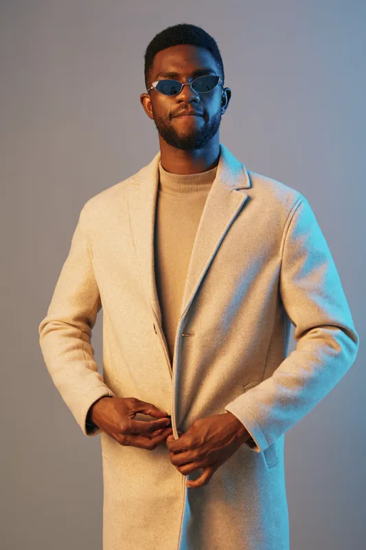 Stylish man in a beige coat and sunglasses, showcasing confidence and elegance against a blue and grey background, ideal for fashion concepts and seasonal trends