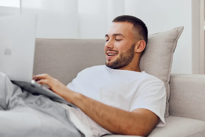 Smiling man relaxing on a sofa, using a laptop with a joyful expression. Modern interior, natural light emphasizes a calm and cozy atmosphere, promoting work-life balance.