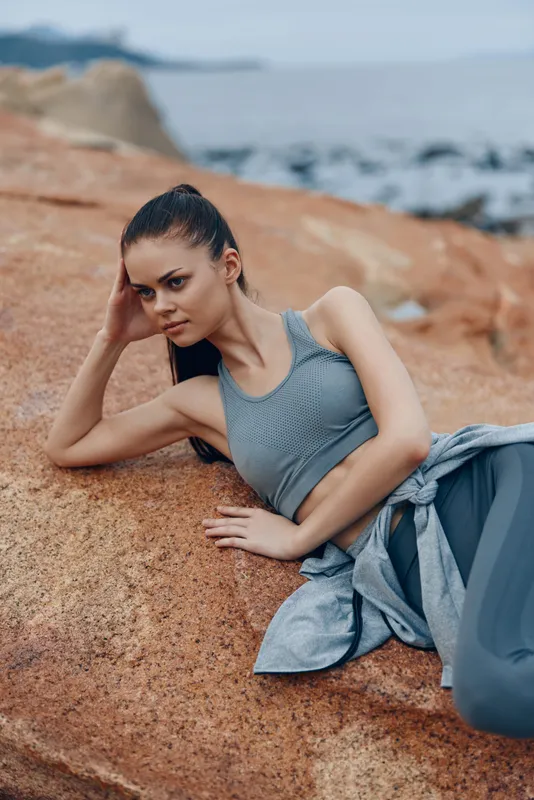 Young athletic woman in stylish workout attire posing on rocky beach, showcasing fitness and wellness lifestyle with serene ocean background