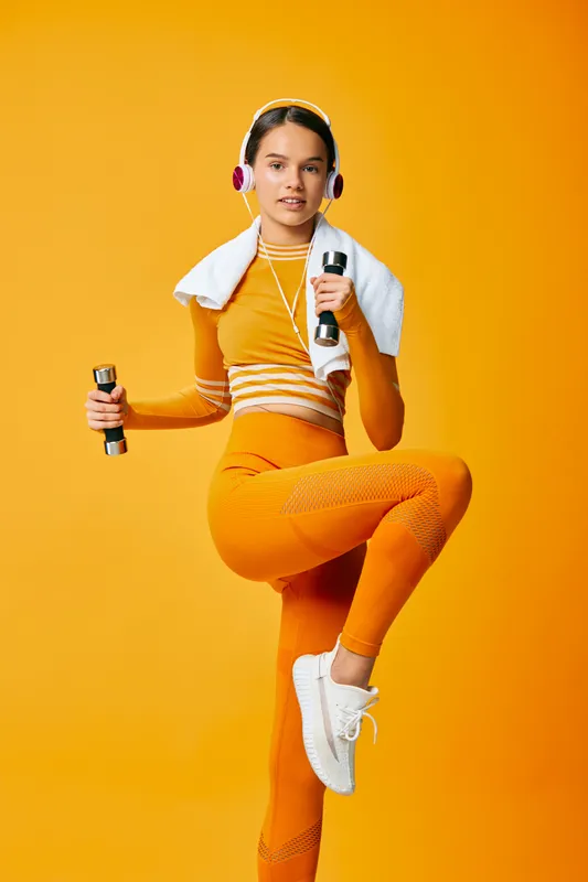 Fitness oriented portrait of a woman in athletic sportwear, dancing pose with a dumbbell and headphones against a vibrant orange background, exuding energy and motivation for workout and exercise.
