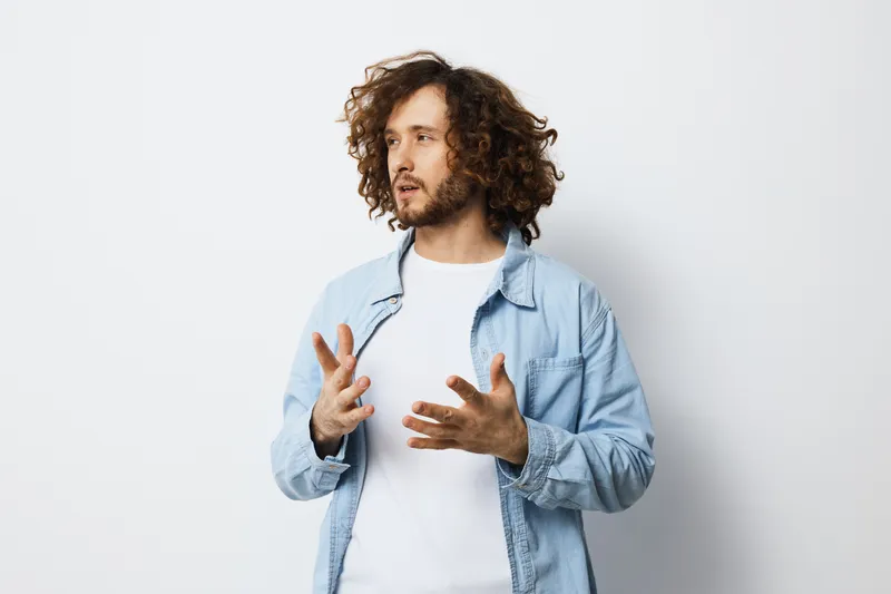 Young man with curly hair expressing emotions while gesturing in a light denim jacket against a plain background Ideal for concepts of communication and self expression