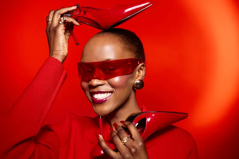 Stylish African woman with dark skin wearing red sunglasses and red sweater holds red high heels near her face on bright red background. Concept for fashion, beauty, and confidence.