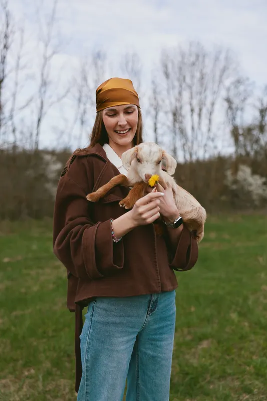 Joyful woman outdoors with a goat kid in her arms, sharing a gentle moment on a grassy field, showcasing lifestyle and care in a natural, warm mood with soft light, style and affection in this casual