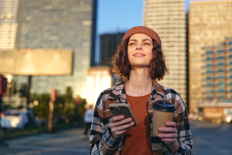 Woman with coffee and phone in urban street, beanie and smile in morning sunlight. Authenticity, candid lifestyle and golden hour glow evoke mindful living and emotional storytelling.
