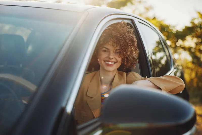 Woman smiling in a car, lifestyle portrait with retro film color, relaxed daylight scene, casual jacket, warm tones, feeling of freedom and joy, natural candid moment