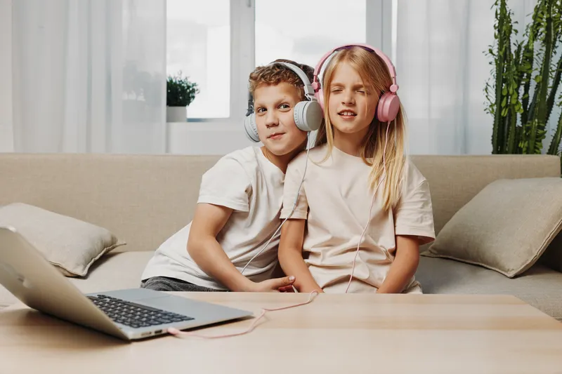 Two children sitting on sofa sharing headphones and listening to music from laptop in bright living room. Concept of friendship, technology, leisure, and modern childhood lifestyle.