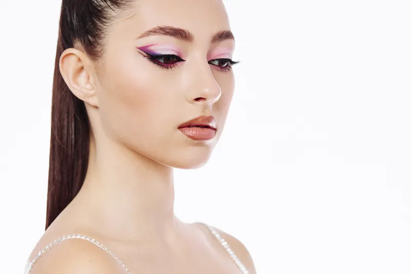 Young woman with creative colorful eyeshadow and sharp eyeliner looks sideways on a white background. The subtle glossy lips and smooth skin highlight beauty and fashion.