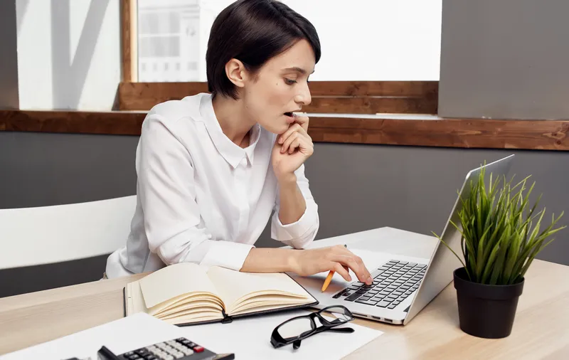 Наследие сидит за столом в офисе, на котором раскрытая тетрадь, цветок в горшке и блокнот
