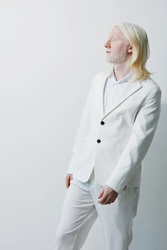 Элегантный мужчина в tailored белом костюме, запечатленный в позе, излучающей уверенность и стиль, на фоне мягкого нейтрального фона, который подчеркивает утонченный внешний вид модели