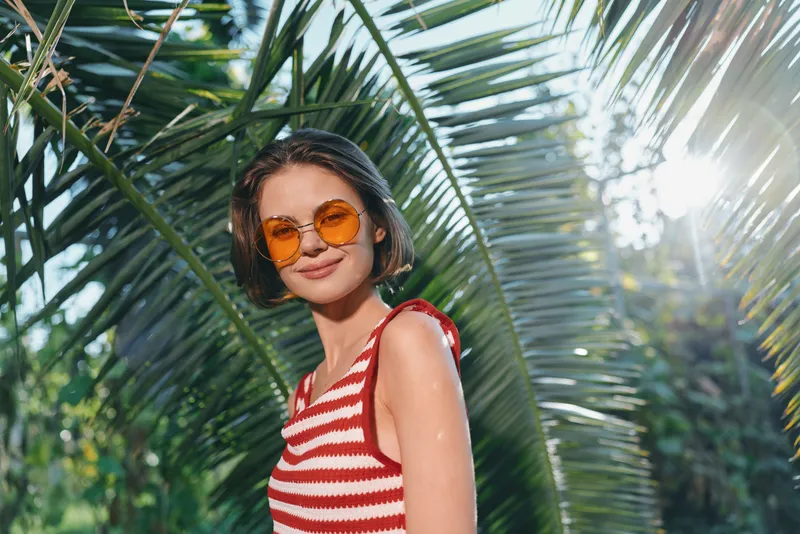 A stylish young woman wearing a red and white striped top and orange sunglasses smiles in a sunny tropical garden, enjoying a relaxed outdoor moment with a carefree vibe.