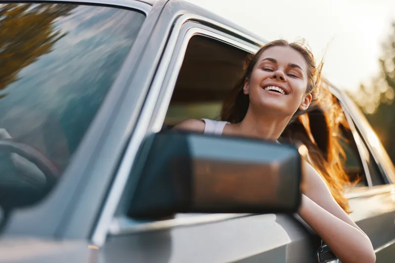 Happy young woman enjoying a summer drive, leaning out of a car window with flowing hair, exuding joy and freedom in a vibrant nature setting.