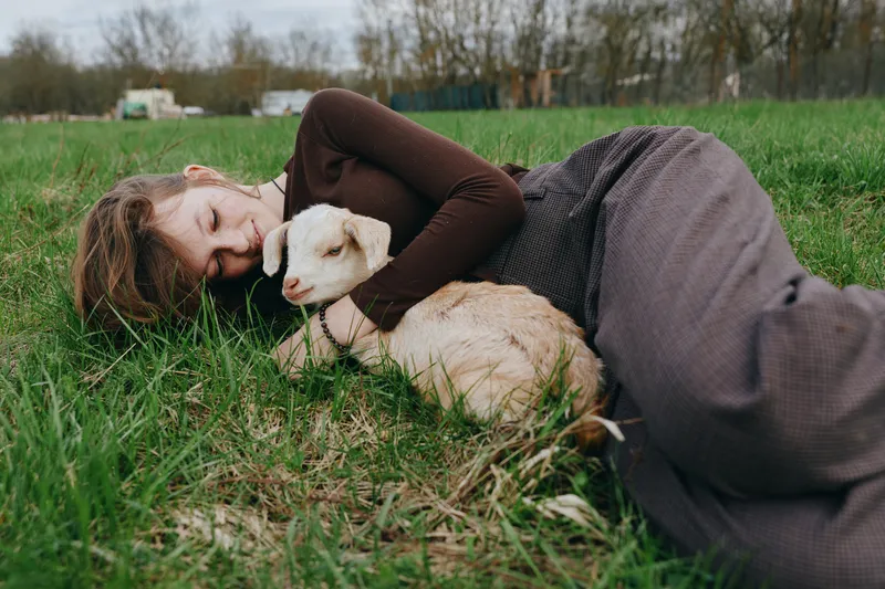 Caring woman lying on grass, embracing a gentle goat kid in a sunny field. Relaxed countryside vibe, companionship, outdoor warmth, wholesome mood, rustic lifestyle.