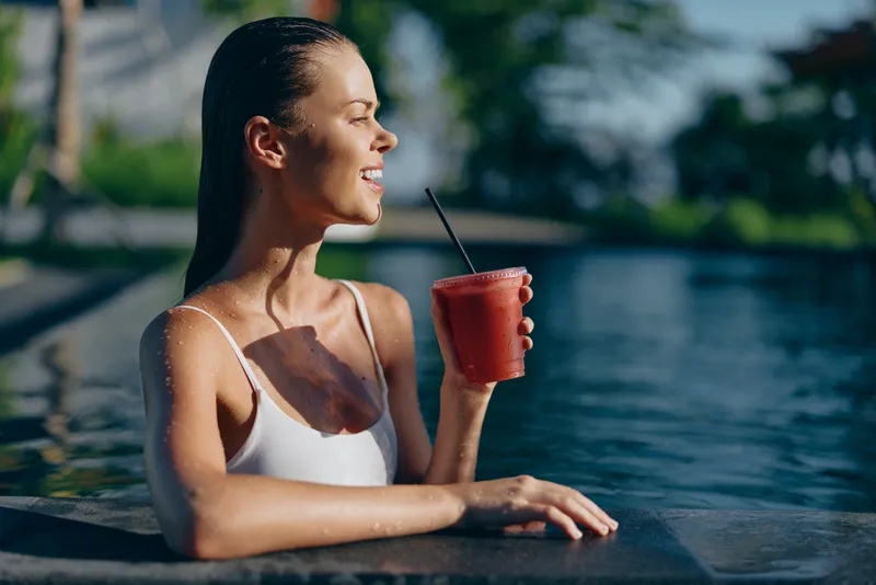 Young woman in a white swimsuit enjoys a refreshing red smoothie by the pool on a sunny day with a relaxed and happy expression.