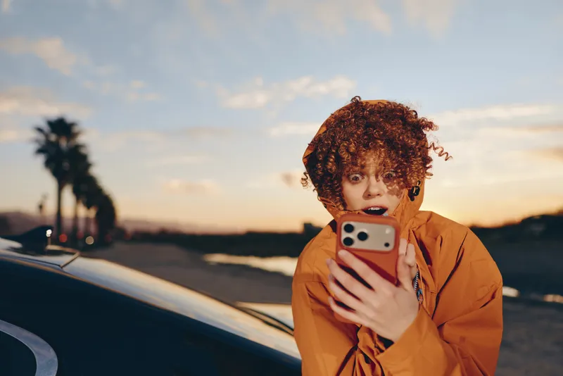 Woman with curly hair holding smartphone for selfie, sunset outdoors near car, casual lifestyle travel mood, warm evening light, candid portrait with soft sky