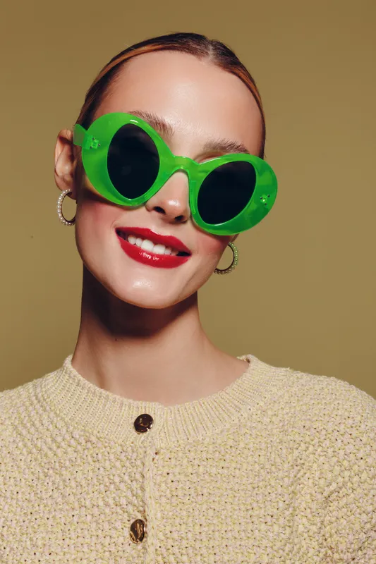 Smiling woman with green oversized sunglasses and red lipstick wears beige knitted sweater and hoop earrings. Studio portrait on neutral background with bright makeup and trendy accessories.