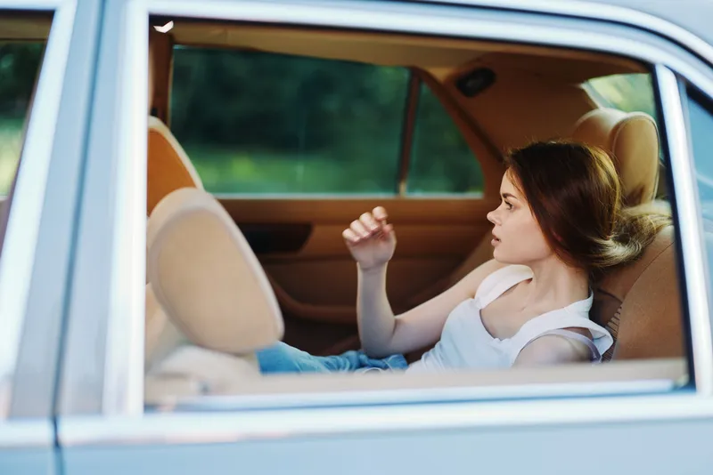 Young woman relaxing in a car, wearing casual clothes, enjoying peaceful moments in nature with soft sunlight illuminating her face and a serene background.