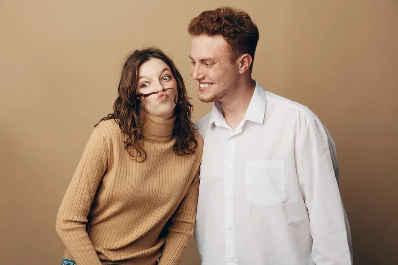 Playful young couple with curly hair and casual clothes making funny faces together on beige background. Concept of friendship, humor, and lighthearted moments between friends.