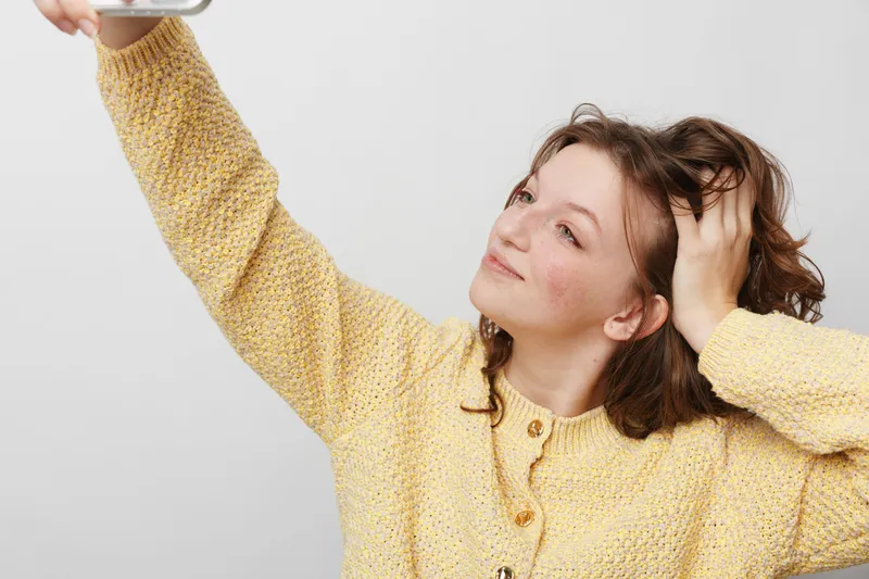 Young woman in yellow knitted sweater takes selfie with smartphone, smiling and touching hair. Casual lifestyle, social media, and self-expression concept on neutral background.