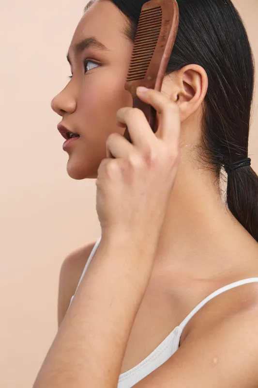 Asian woman styling her hair with a wide wooden comb in a clean studio setting. Calm expression, focused moment, natural light highlighting smooth skin and quiet elegance.