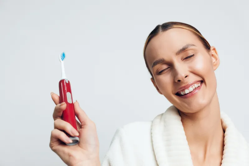 Young woman with blonde hair smiles with closed eyes while holding a red electric toothbrush against a plain light background.