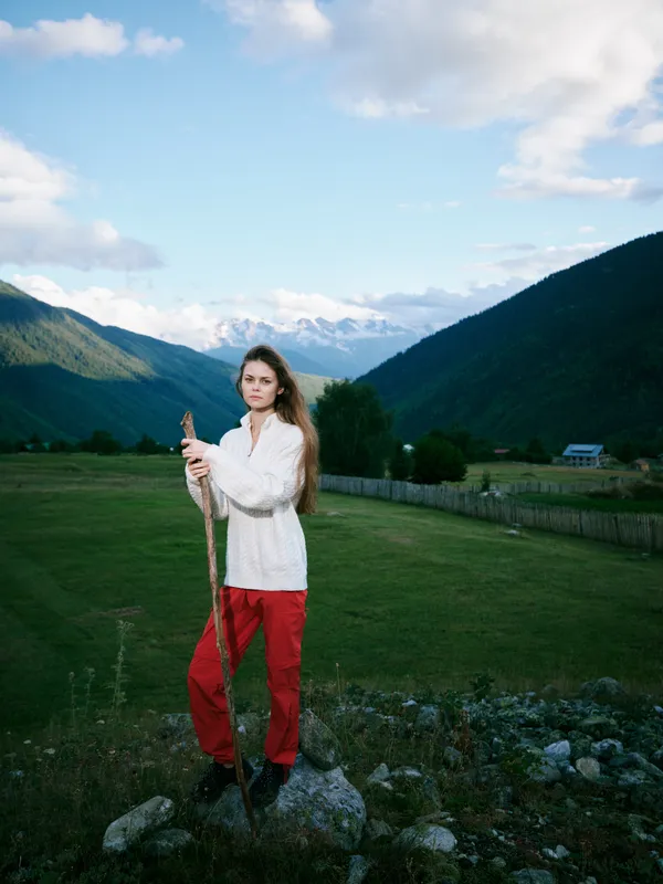 Mountain field outdoor scene with a woman in red pants holding a staff, valley views, clear sky and rustic fence, capturing nature and exploration in wide rural landscape