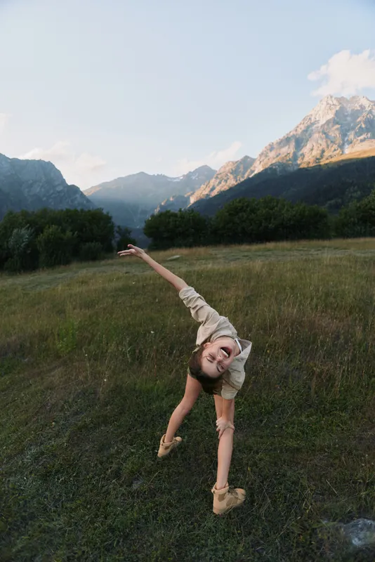 A young woman in a light beige outfit leaps joyfully in a mountain meadow, arms outstretched, with towering peaks in the background, conveying carefree outdoor play.