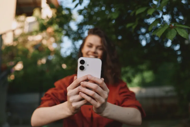 Smiling woman taking a selfie outdoors with a smartphone. Casual outfit, warm natural light, emphasizes lifestyle, self-expression, and connectivity
