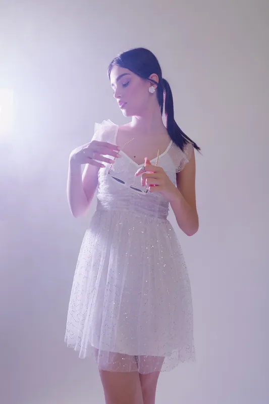 Young woman in a white sparkling dress holds a glass and a cigarette, posing thoughtfully in soft studio lighting. Concept for beauty, fashion, and elegance.