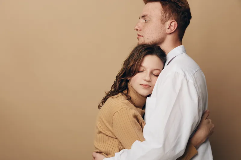 Young couple embraces tenderly with closed eyes, expressing love and comfort. Woman in brown sweater hugs man in white shirt against beige background. Concept of affection and intimacy.