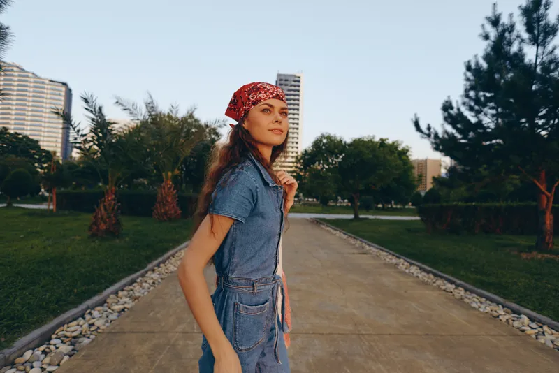 young woman walking outdoors in denim dress and red headscarf on sunny day with urban buildings and green trees in background