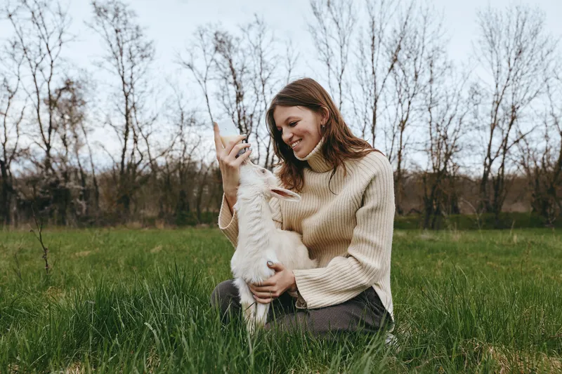 A smiling woman gently comforts a young goat in a sunlit field, kneeling among tall grass. A warm countryside lifestyle moment, highlighting bonding between human and animal outdoors, in nature.