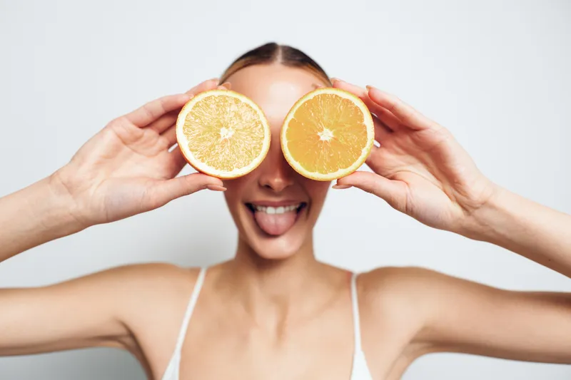 Young woman with long blonde hair playfully holds two halves of an orange in front of her eyes while sticking out her tongue against a plain white background.